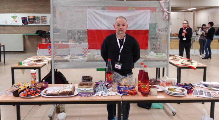Pan Łukasz Piotrowski stojący za stołem z poczęstunkiem. W tle polska flaga.