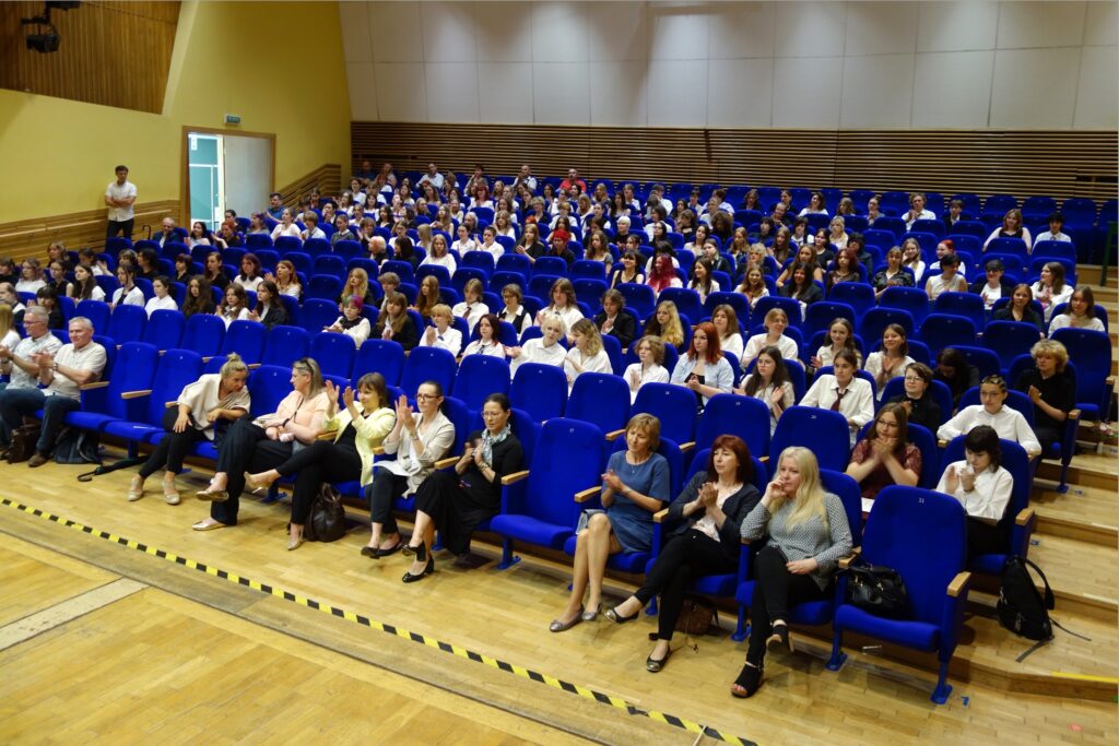 Nauczyciele i uczniowie siedzący na widowni na zakończeniu roku szkolnego.